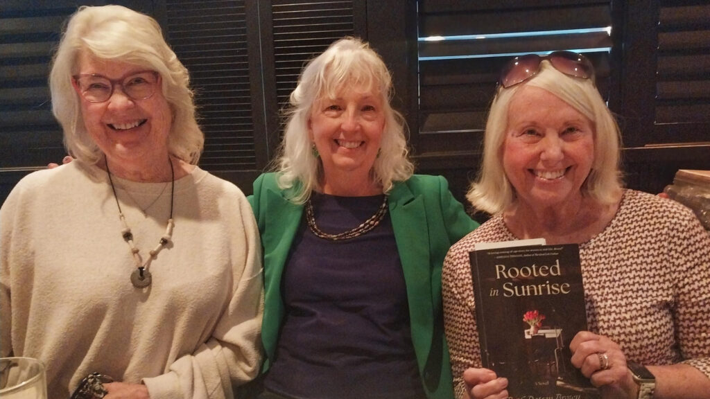 Three women smiling at camera while holding up a copy of Rooted in Sunrise